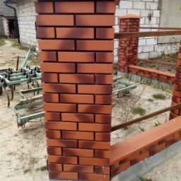 Fragment ogrodzenia z brązowej cegły klinkierowej z ciemniejszymi przebarwieniami, z metalową, rdzawą poręczą, na tle białego budynku z numerem 19a oraz zielonego sprzętu rolniczego.