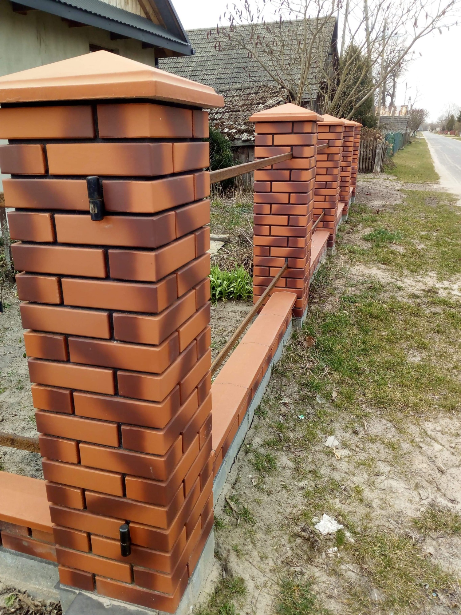 Nowoczesne ogrodzenie z cegły klinkierowej z metalowymi przęsłami, widoczne słupki i podmurówka, otoczenie trawnik z widocznymi śladami po budowie.
