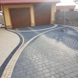 Ułożona kostka brukowa w dwóch odcieniach szarości z dekoracyjnymi wstawkami w kolorze beżowym, tworząca wzór na podjeździe przed garażem z brązowymi drzwiami.