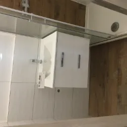 Biała szafka pod umywalką z chromowanymi uchwytami, zamontowana na ścianie z białych płytek w łazience z drewnopodobną podłogą, widoczny fragment szklanej kabiny prysznicowej.