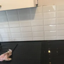 Białe, wypukłe płytki na ścianie nad czarnym, błyszczącym blatem kuchennym ze zlewozmywakiem, gąbką i ścierką