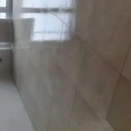 Beżowe płytki ułożone diagonalnie na podłodze, widoczne odbicie światła od okna, fragment ściany z otworem rewizyjnym.