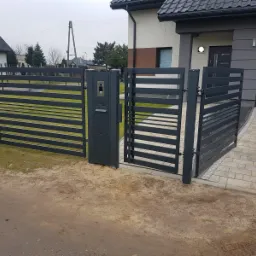 Nowoczesna, grafitowa brama wjazdowa z poziomymi panelami, skrzynką na listy i domofonem, prowadząca na posesję z kostki brukowej, na tle domu z szarą elewacją i garażem.