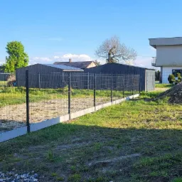 Montaż ogrodzenia panelowego 3D z podmurówką betonową, widoczne słupki i panele w kolorze grafitowym, na trawiastym terenie z budynkami w tle.