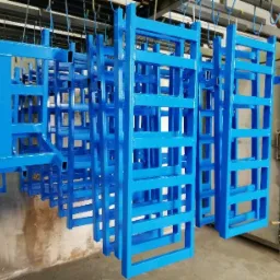 Metalowe konstrukcje regałowe, pomalowane na niebiesko, zawieszone na hakach wewnątrz lakierni przed wygrzewaniem.