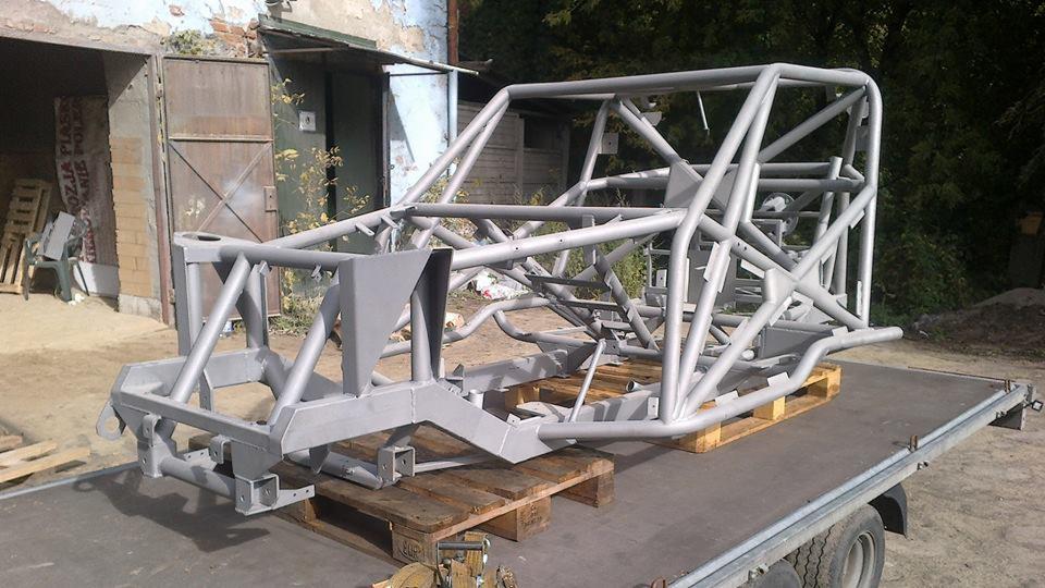 Rama pojazdu terenowego po obróbce, pomalowana na szaro, umieszczona na paletach na przyczepie, widoczne ślady korozji na ścianie budynku w tle.