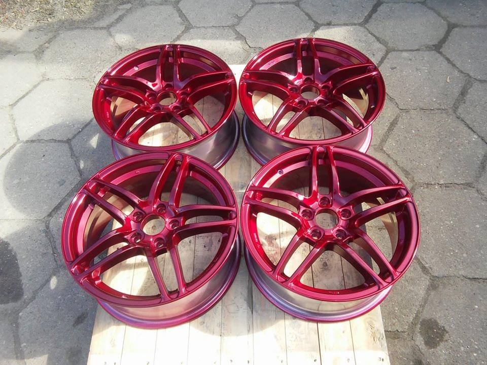 Cztery felgi samochodowe po malowaniu proszkowym na intensywny, metaliczny odcień czerwieni, ułożone na palecie drewnianej na tle szarej kostki brukowej.