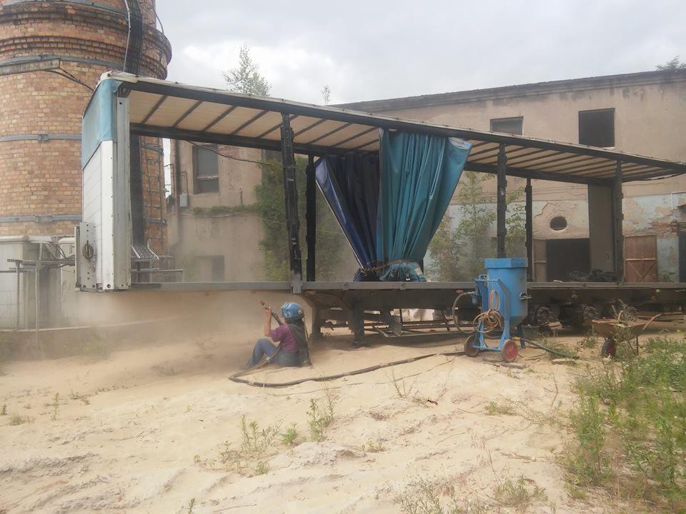 Pracownik w kasku ochronnym, siedząc na piaszczystym podłożu, wykonuje czyszczenie strumieniowo-ścierne konstrukcji z użyciem mobilnego urządzenia, na tle ceglanego komina i zniszczonego budynku.