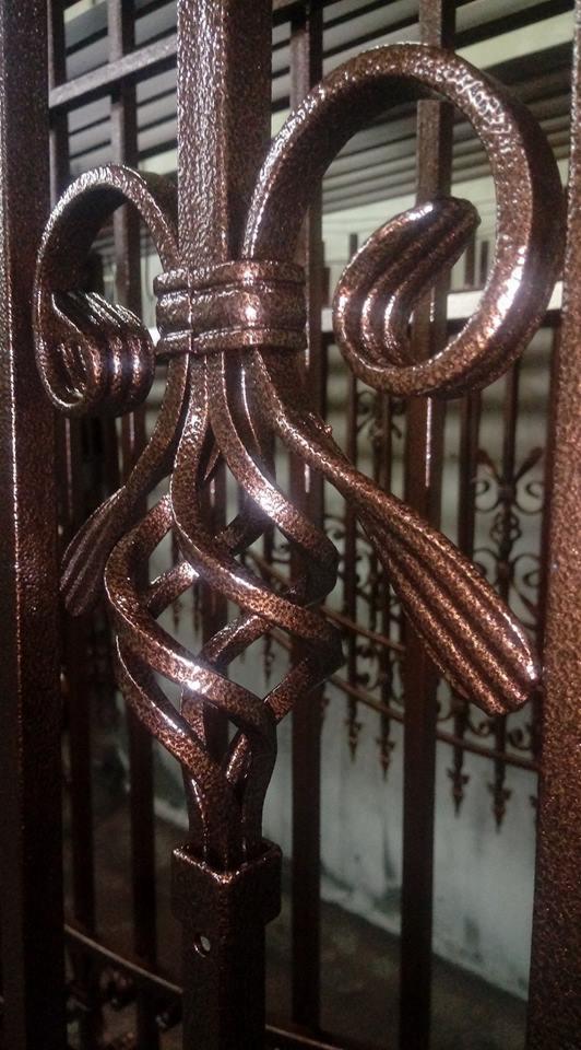 Dekoracyjny element metalowy ogrodzenia, pomalowany na brązowo z efektem młotkowym, z widoczną teksturą i spiralnymi zdobieniami.