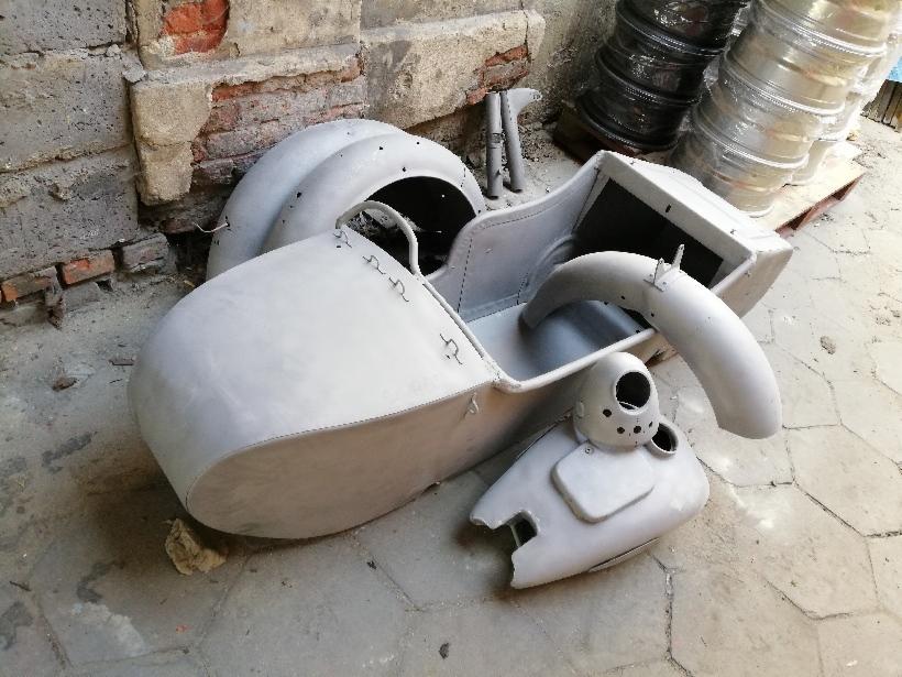 Elementy motocykla (boczny wózek, błotnik, zbiornik) po obróbce przygotowującej do malowania, leżące na podłodze w pobliżu ceglanej ściany.