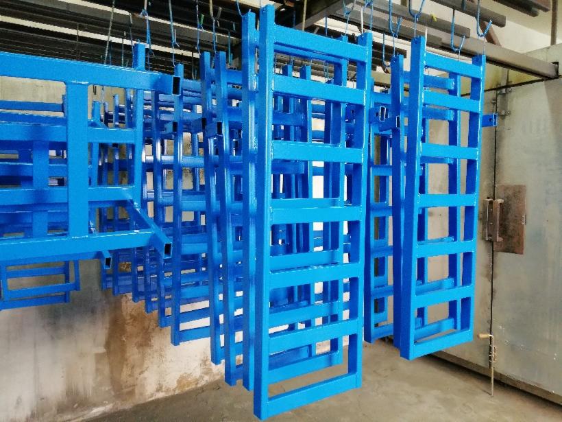 Metalowe konstrukcje regałowe, pomalowane na niebiesko, zawieszone na hakach wewnątrz lakierni przed wygrzewaniem.