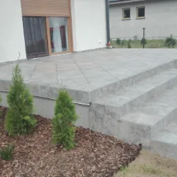 Wykończony taras z szarych płytek ceramicznych i schodami, obok posadzone małe tuje, widok na budynek z roletami zewnętrznymi.