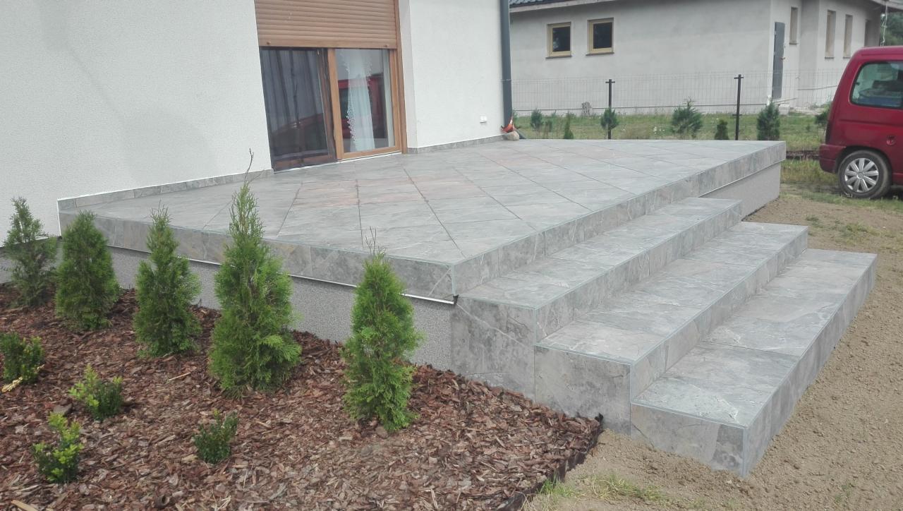 Wykończony taras z szarych płytek ceramicznych i schodami, obok posadzone małe tuje, widok na budynek z roletami zewnętrznymi.