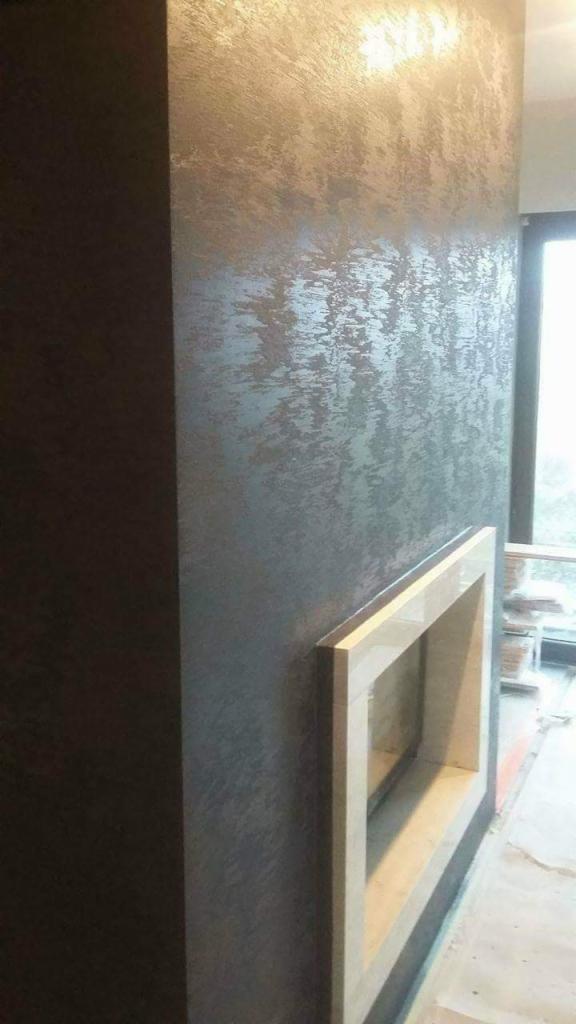 Dekoracyjna ściana z efektem metalicznego przetarcia w odcieniach szarości, z widocznym fragmentem obudowy kominka w trakcie montażu.