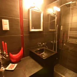 Ciemna łazienka z akcentami czerwieni: świece na srebrnym świeczniku, dekoracyjna misa, kwadratowe lustro w złotej ramie, czarna umywalka i grzejnik drabinkowy w kabinie prysznicowej.