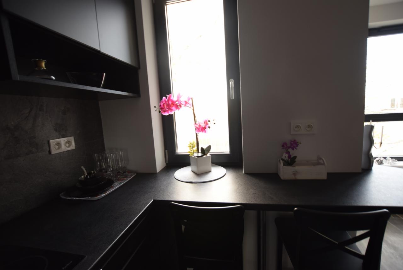 Ciemna kuchnia z czarnymi szafkami, blatem i krzesłami, ożywiona różowymi kwiatami w doniczce na tle okna i drugą małą doniczką z fioletowymi kwiatami w ozdobnej skrzynce.