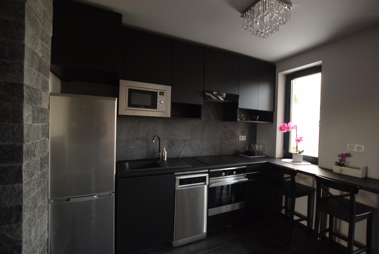 Nowoczesna kuchnia w czerni i szarości z elementami stali nierdzewnej, kryształowym żyrandolem, kwiatami w oknie i ścianą wykończoną kamieniem łupkowym.