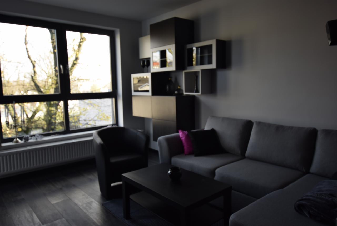 Nowoczesny salon w odcieniach szarości z akcentem fuksji na poduszce, minimalistyczna zabudowa meblowa z podświetlanymi półkami, czarny skórzany fotel obok okna z widokiem na drzewa.