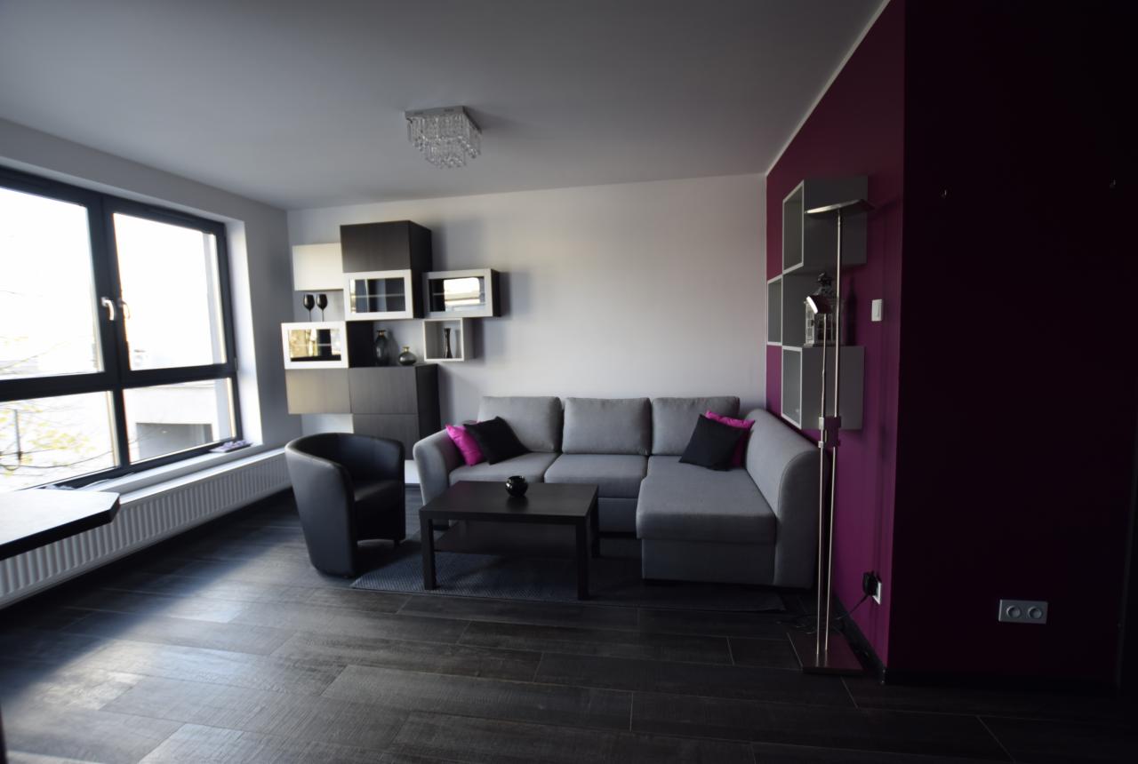 Nowoczesny salon z szarą narożną sofą, czarnym stolikiem kawowym i fotel, ciemna podłoga imitująca drewno, fioletowa ściana akcentująca z półkami, dekoracyjny regał wiszący na ścianie...