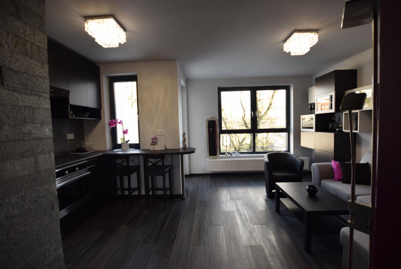 Ciemna aranżacja wnętrza mieszkania: kuchnia z barkiem i salon z szarą sofą, czarnym fotelem i stolikiem kawowym, podłoga z ciemnego drewna, dwa ozdobne żyrandole.