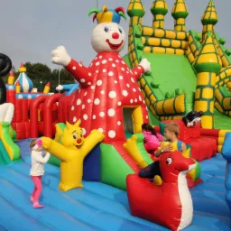 Dzieci bawią się na dmuchanym placu zabaw z motywami postaci z bajek, w tym dmuchany klaun w czerwonym stroju w białe kropki, żółty lew z podniesionymi rękami i zamek z żółtymi wieżami.