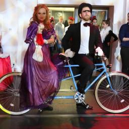 IN SPE grupa marketingowa - Para w kostiumach na tandemie Cinelli pozuje na tle gości i bufetu z napojami podczas tematycznego wydarzenia.