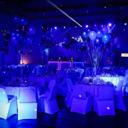 IN SPE grupa marketingowa - Uroczysta sala balowa oświetlona intensywnym niebieskim światłem, z okrągłymi stołami nakrytymi białymi obrusami i krzesłami z białymi pokrowcami, dekoracje z balonów w odcieniach niebieskiego...