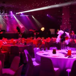 IN SPE grupa marketingowa - Uroczysta sala bankietowa z okrągłymi stołami udekorowanymi białymi obrusami, szklanymi dzbankami z pomarańczowym napojem i piórami w wazonach, oświetlona reflektorami w kolorach różu i fioletu,...