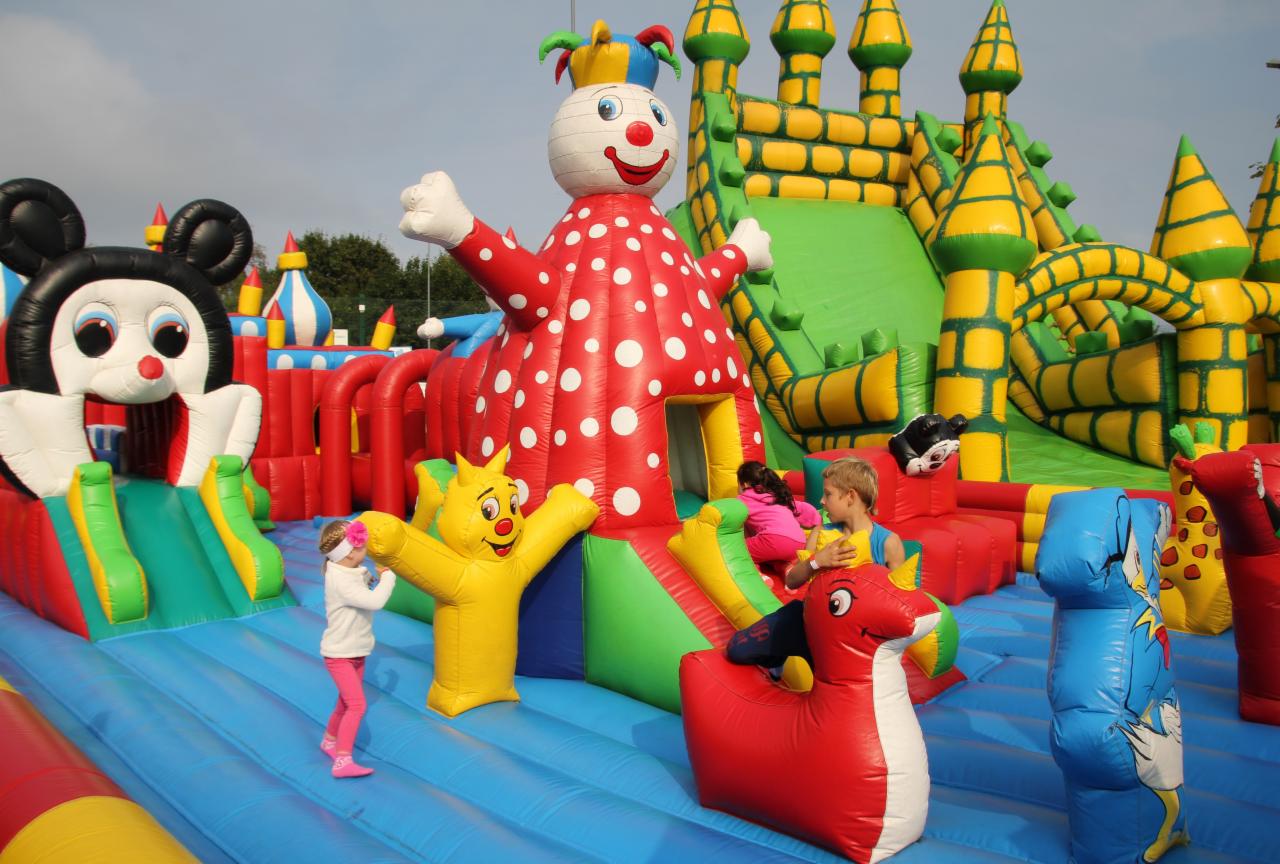 Dzieci bawią się na dmuchanym placu zabaw z motywami postaci z bajek, w tym dmuchany klaun w czerwonym stroju w białe kropki, żółty lew z podniesionymi rękami i zamek z żółtymi wieżami.