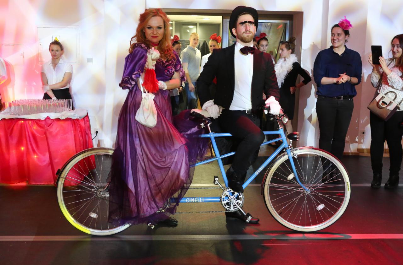 Para w kostiumach na tandemie Cinelli pozuje na tle gości i bufetu z napojami podczas tematycznego wydarzenia.