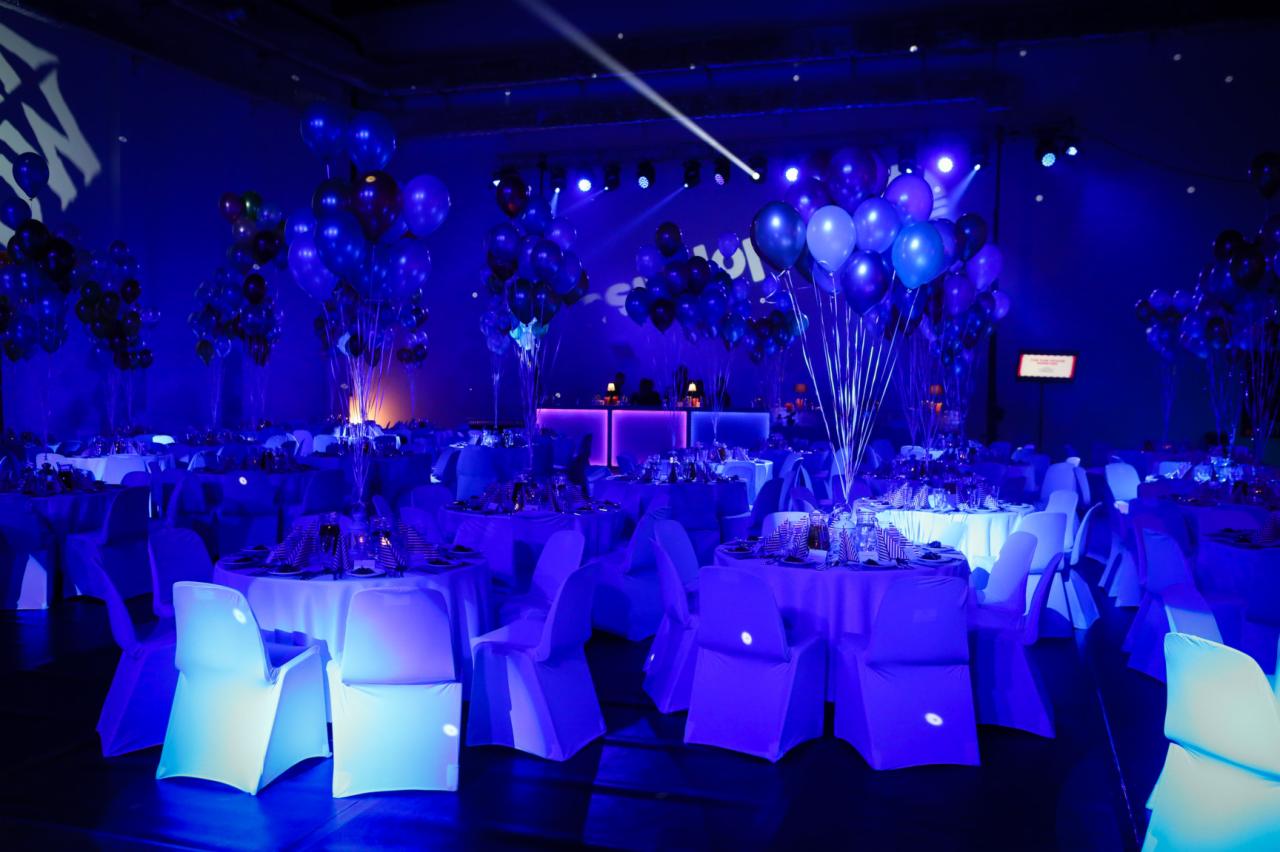 Uroczysta sala balowa oświetlona intensywnym niebieskim światłem, z okrągłymi stołami nakrytymi białymi obrusami i krzesłami z białymi pokrowcami, dekoracje z balonów w odcieniach niebieskiego...