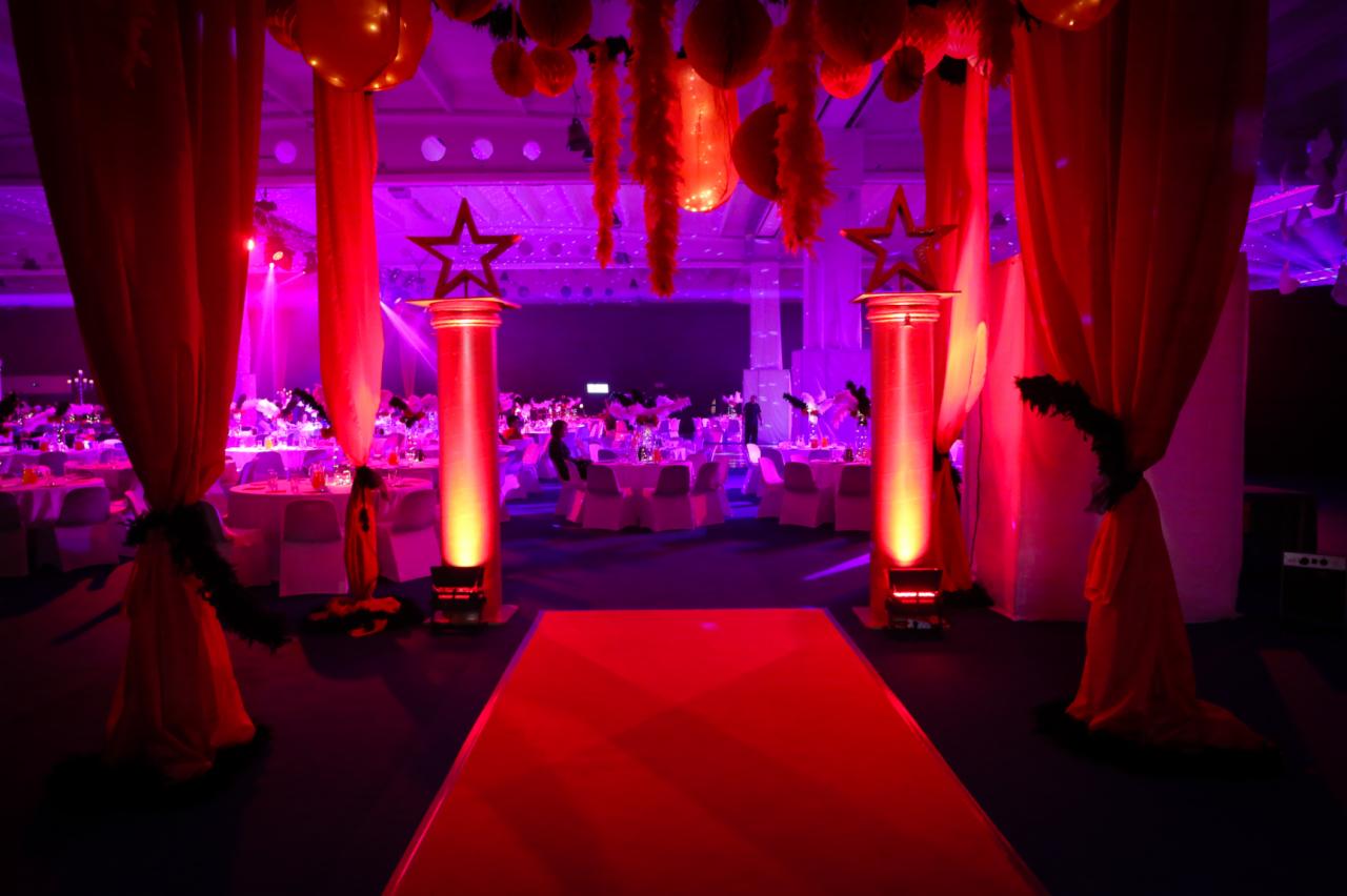 Uroczysta sala bankietowa oświetlona fioletowym światłem, z czerwonym dywanem prowadzącym przez środek, kolumny z gwiazdami, wiszące ozdoby i elegancko nakryte stoły.