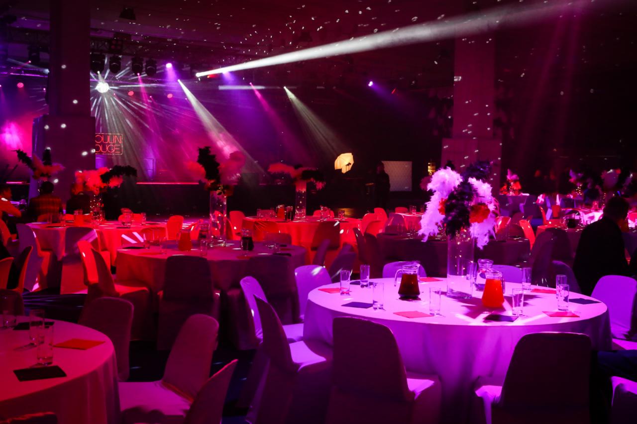 Uroczysta sala bankietowa z okrągłymi stołami udekorowanymi białymi obrusami, szklanymi dzbankami z pomarańczowym napojem i piórami w wazonach, oświetlona reflektorami w kolorach różu i fioletu,...