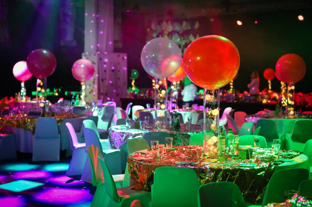 Uroczysta sala balowa z okrągłymi stołami nakrytymi błyszczącymi obrusami, krzesła w odcieniach zieleni i błękitu, dekoracje z balonów wypełnionych światełkami, oświetlenie sceniczne w kolorach...