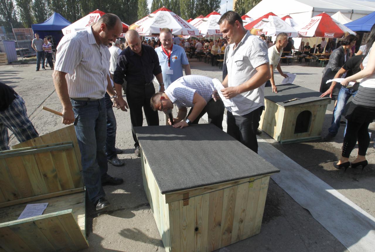 Grupa mężczyzn ocenia budę dla psa z drewnianych desek i bitumicznym dachem podczas plenerowego konkursu, widoczne parasole Tyskie w tle.