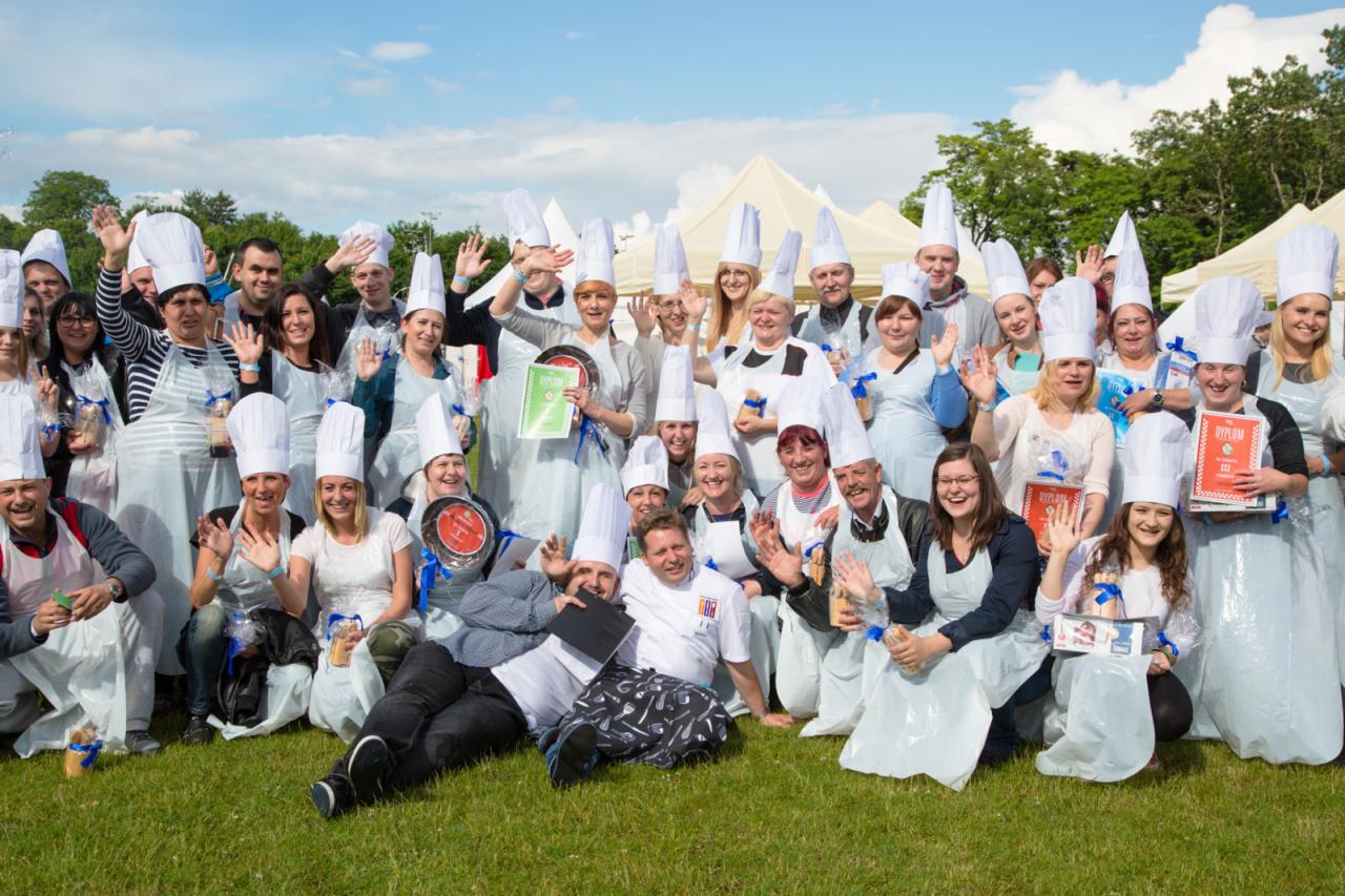 Duża grupa uśmiechniętych osób w białych czapkach kucharskich i plastikowych fartuchach pozuje na trawie, niektórzy trzymają dyplomy i małe upominki, w tle białe namioty i drzewa.
