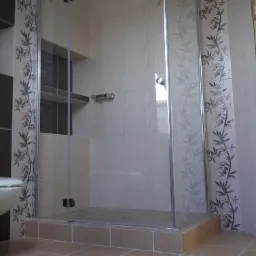 Kabina prysznicowa ze szklanymi drzwiami i płytkami z motywem roślinnym, zamontowana w łazience z beżowymi płytkami podłogowymi i ściennymi. Widoczny fragment toalety po lewej stronie.