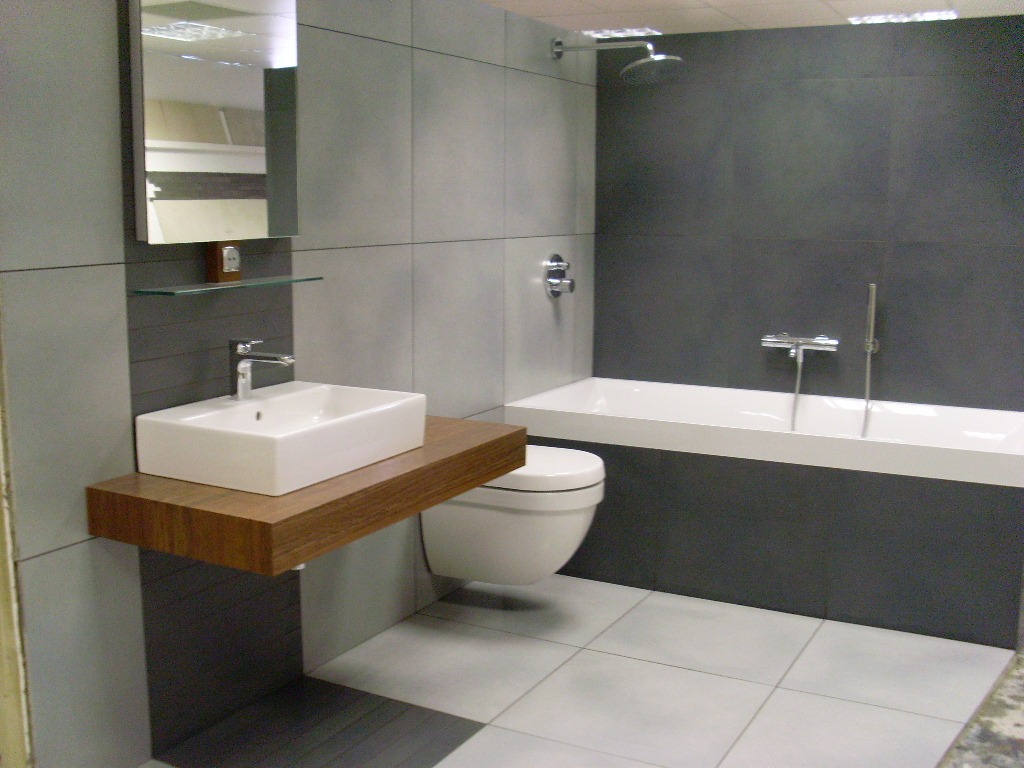 Nowoczesna łazienka z szarymi płytkami, białą umywalką na drewnianym blacie, toaletą i wanną z prysznicem.