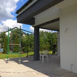 Biała elewacja domu z szarymi elementami wykończeniowymi, widoczne rusztowanie, pracownik w tle, plastikowe krzesło i ślady prac budowlanych na podłożu.