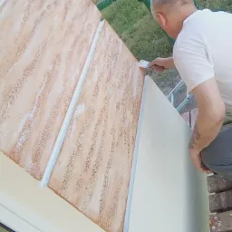 Pracownik z tatuażem na ramieniu, w białej koszulce i szarych spodniach, aplikuje pędzlem białą farbę na fragment elewacji z dekoracyjną, brązowo-białą fakturą, praca w toku.