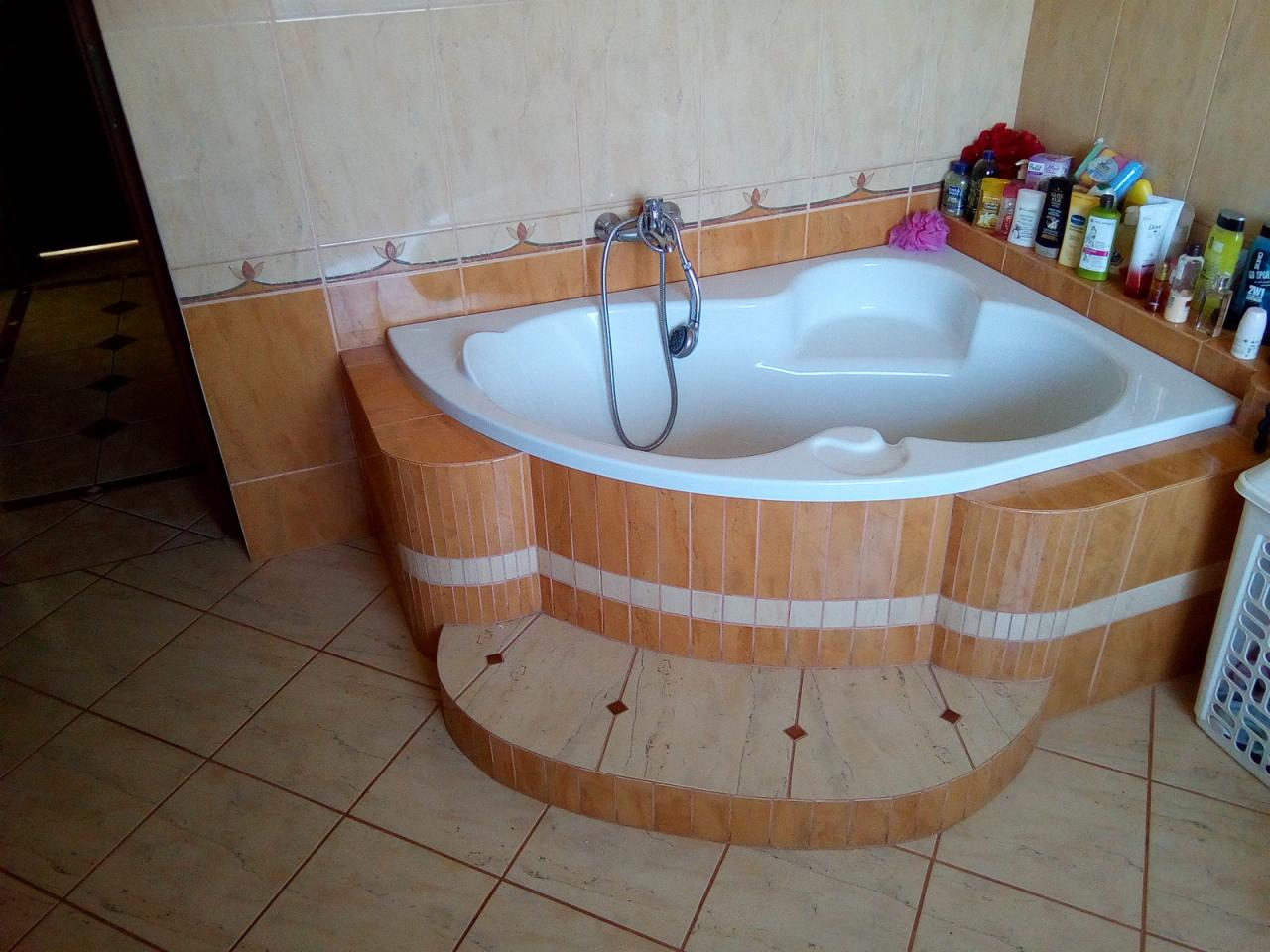 Narożna wanna z hydromasażem, obudowana mozaiką w odcieniach beżu i brązu, z widoczną armaturą i półką z kosmetykami w tle, ustawiona na podwyższeniu z płytek.