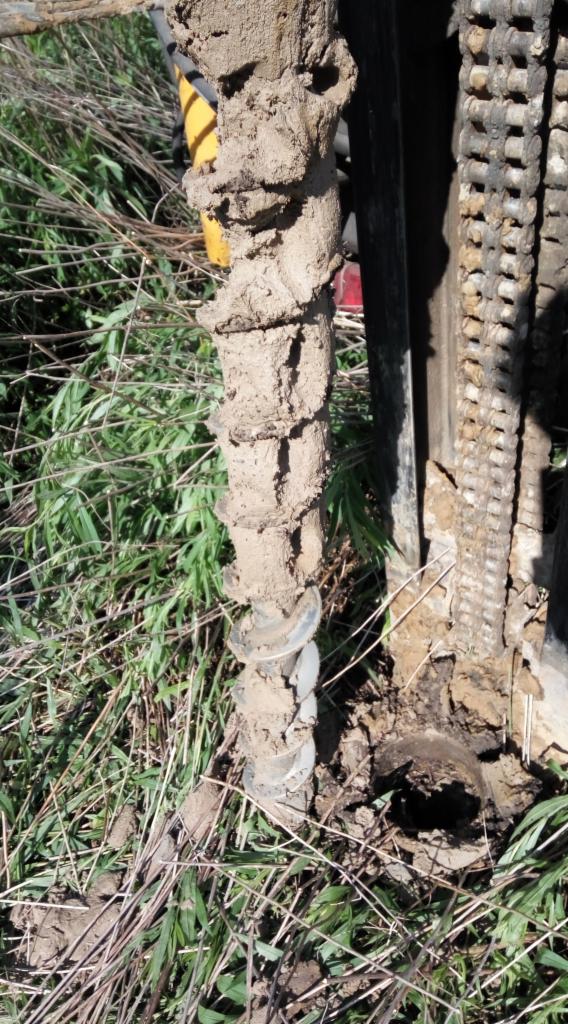 Wiertło świdrowe pokryte błotem, w trakcie wiercenia otworu w ziemi porośniętej trawą.