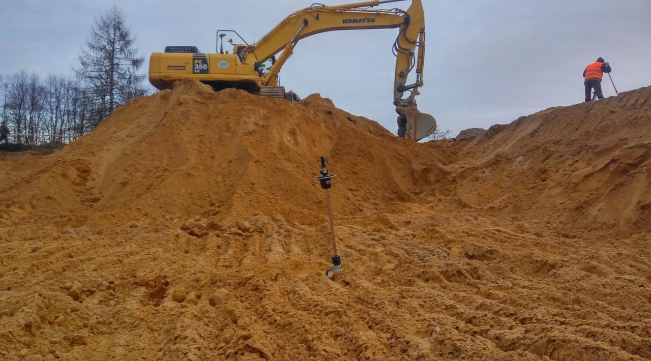 Żółta koparka Komatsu na szczycie piaszczystego wzgórza, geodeta z tyczką w pomarańczowej kamizelce, w tle drzewa bez liści i pochmurne niebo.