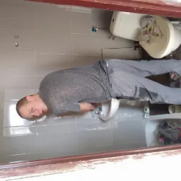 Mężczyzna w szarych spodniach i koszulce instaluje białą umywalkę w łazience z szarymi płytkami, widoczne narzędzia i części instalacyjne, fragment toalety w tle.