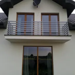 Szara, pozioma balustrada balkonowa z profili zamkniętych na tle elewacji domu z brązowymi oknami i ciemnym dachem.