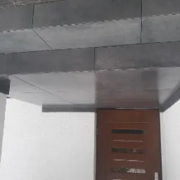 Zadaszenie wejścia do budynku wykończone ciemnoszarymi płytkami ceramicznymi, widoczna brązowa stolarka drzwiowa.