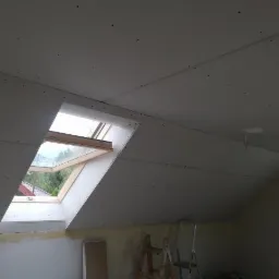 Montaż płyt kartonowo-gipsowych na poddaszu z oknem dachowym, widoczne szpachlowane łączenia i drabina oparta o ścianę.