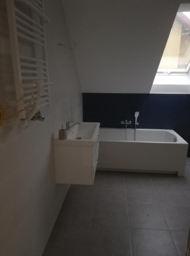 Nowoczesna łazienka w odcieniach bieli i szarości z wanną, umywalką z szafką i grzejnikiem drabinkowym, oświetlona przez okno dachowe.