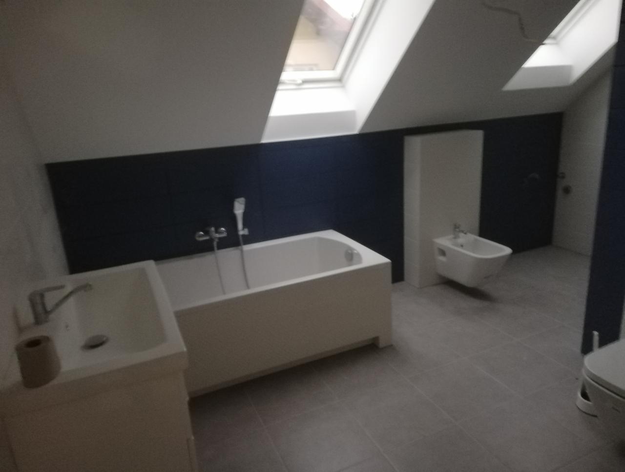 Nowoczesna łazienka na poddaszu z wanną, bidetem i umywalką, wykończona w odcieniach bieli i granatu, oświetlona światłem z okien dachowych.