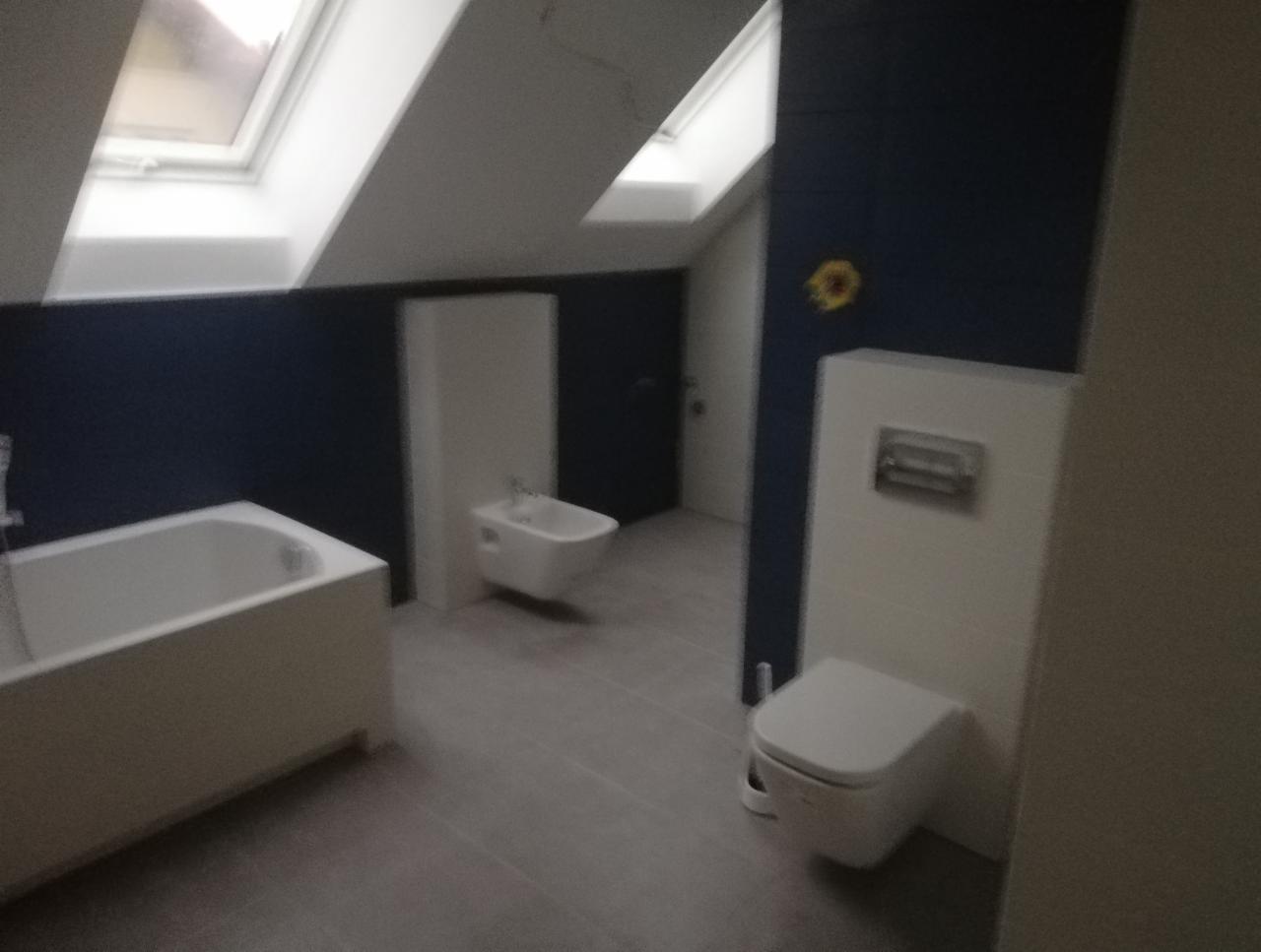 Nowoczesna łazienka na poddaszu z wanną, bidetem i toaletą w zabudowie, wykończona ciemnoniebieskimi panelami ściennymi i szarą podłogą, oświetlona przez okna dachowe.